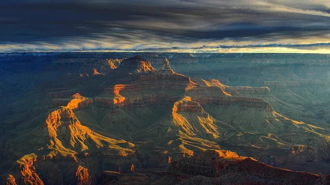 Yellow sunrise light just above the horizon striking sideways onto peaks and ridges with within Grand Canyon. A thick layer of dark clouds is overhead.