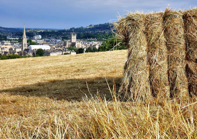A photo of a freshly cut hayfield, with a hay bale in the foreground and the spires and towers of Bath city centre in the background.