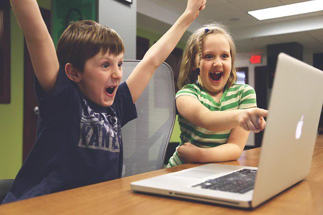 A boy sits in front of a computer with his arms raised above his head. A girl points at the screen and smiles.