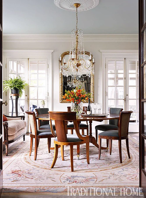 A bright dining room is filled with sunlight from large french doors at the back of the room. A beautiful crystal chandelier hangs above a round antique dining table set for six people. A velvet settee sits next to the table.