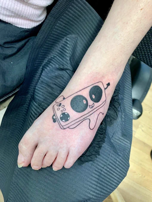 Ein Tattoo vom Xbox Adaptive Controller. Im Comic-Stil niedlich gezeichnet mit den Knöpfen als Augen und kleinen Ärmchen.