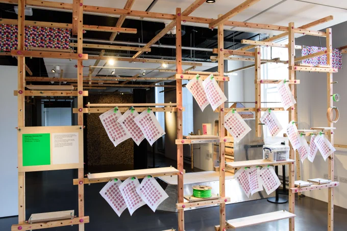 A wooden structure made of thin wooden posts, connected by pink screws. A4 sheets of fabric printed with pink circles hang off the horizontal beams, while a patterned felt wall of another artwork can be seen in the background through the shelf spaces