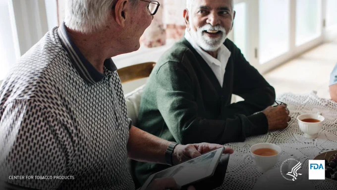 Two men at a table having tea. One man is holding an iPad and they are looking at each other and smiling. 