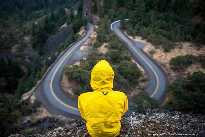Person in yellow rain jacket looking down from a hilltop onto a switchback road. Image credit: Free-photos via Pixabay.