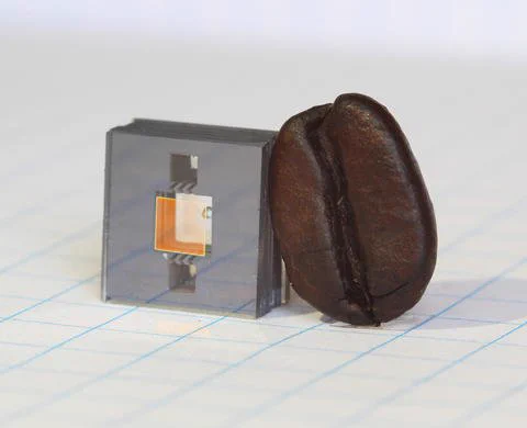 Square gray cube with two square holes in it and a gold square in between the holes, and a dark brown coffee bean leaning against the cube.