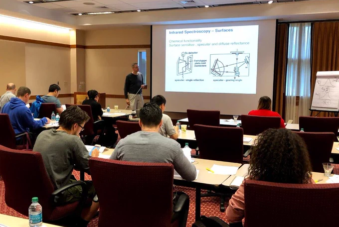 Professor stands at front of room discussing a powerpoint slide as students watch.