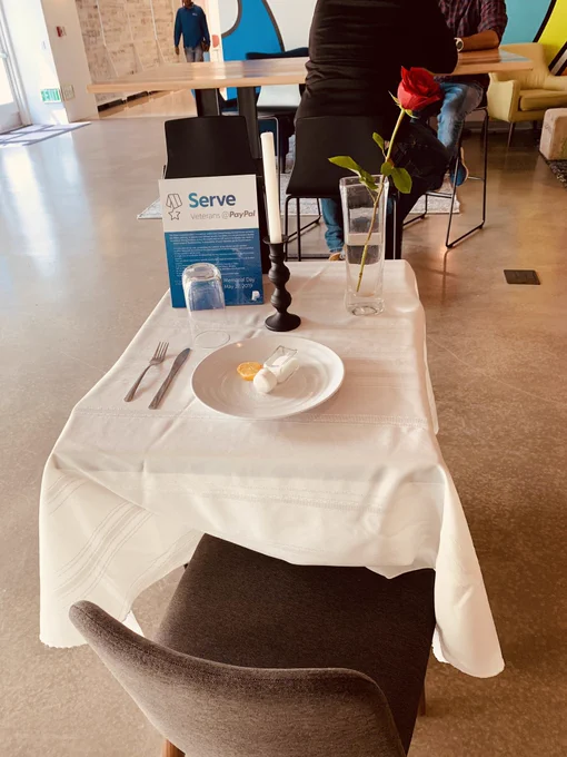 Small table with one single chair, white table cloth, white dinner plate with a lemon slice and salt shaker, one glass inverted, a glass with a single red rose, one single white candle.