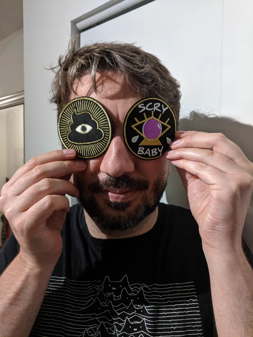 Me holding two patches over my eyes. Both are gold on black, and each has a depiction of an eye.