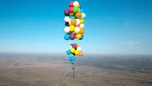 Un hombre vuela sustentado por docenas de globos de colores, en un día de cielo claro y celeste.
