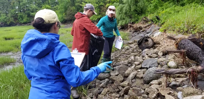 one person putting trash in garbage bag and holding clipboard, one man holding trash bag and one woman with back facing holding clipboard