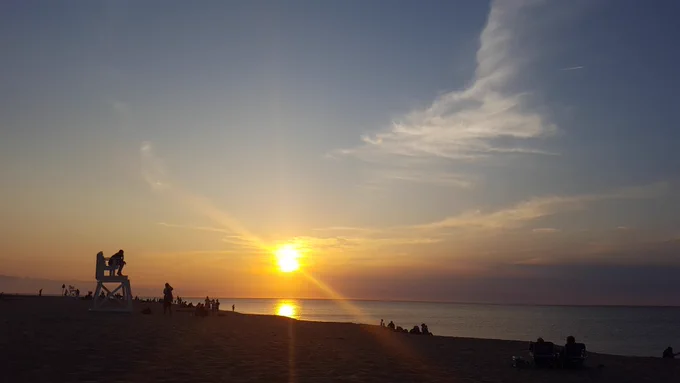 The silhouettes of a large group of people on the beach are highlighted by a swirling golden sunset behind wispy clouds over a calm ocean.