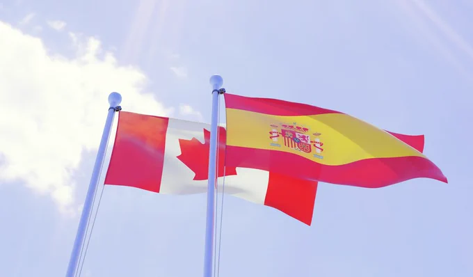 The Canadian flag flying behind the Spanish flag under a blue sky.