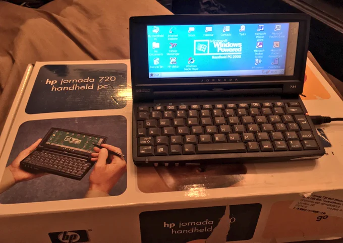 A handheld PC from the 2000s sits on top of its box that says HP Jornada 720 Handheld PC. The Windows CE desktop is displayed.