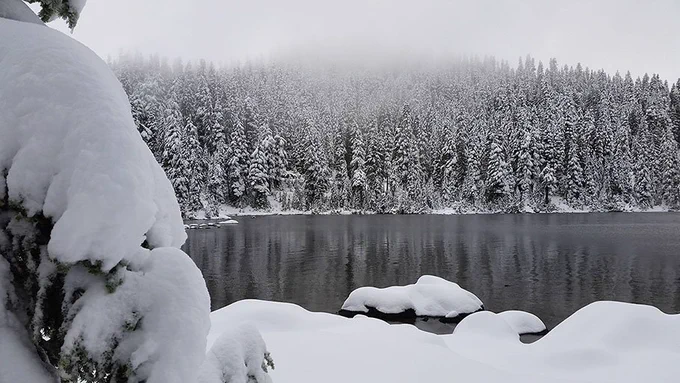Snow covered trees and rocks surround a lake.