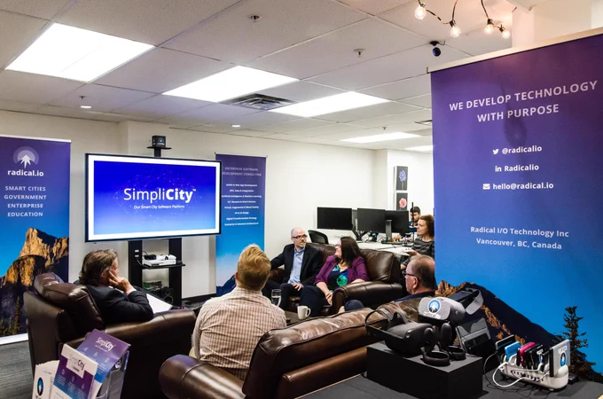 A group of people are gathered in a meeting room looking at a wall-mounted television with "SimpliCity - Our Smart City Software Platform" displayed. 