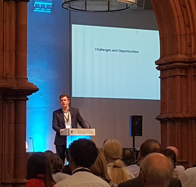 Man at podium with screen behind and red stone pillars either side