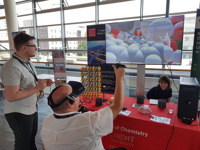 Person sat down wearing a VR headset in front of a screen behind a table. Person stood to their left looking at screen.