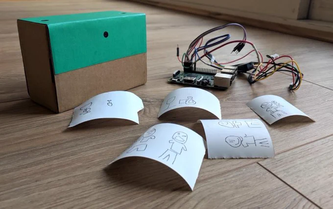 A small cardboad box with a hole for a camera lens, a Raspberry Pi Model 3B with a tangle of jumper leads connected, and five pieces of thermal printer paper, each bearing rough doodles of people, plants, and everyday objects
