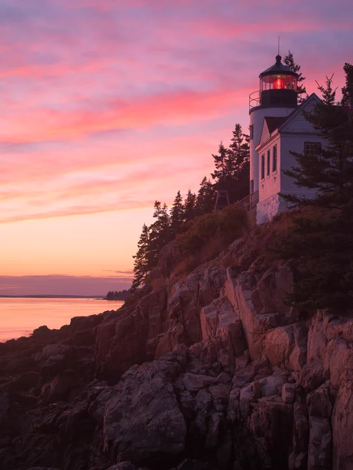 A small white building with a lighthouse tower stands on a rocky cliff overlooking a bay at sunset.