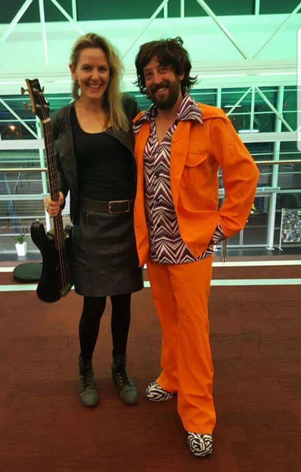 Two people in fancy dress smiling at the camera. A tall woman is in black leather holding a guitar and a man is wearing a dark wig and bright orange jumpsuit with zebra print high heeled shoes.