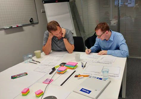 Two men, sitting at a table with lots of paper, pens and post-it notes, sketching