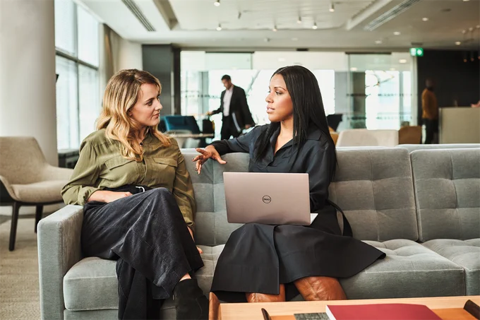 Two females sitting on a Sofa in an office environment. One is using a Surface book.