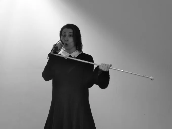 B&W -- a young woman in black holds and partly folds a white cane, pretending it's a microphone. She pouts.