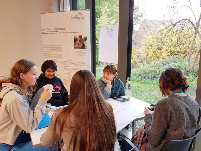 5 Teilnehmende am Tisch diskutieren. Hintergrund Plakat von Verein bridge it .