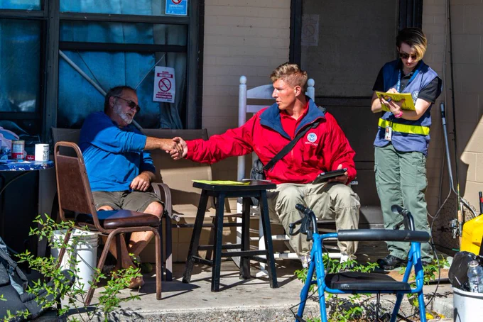 A FEMA employee sits and shakes the hand of a disaster survivor on the porch of his damaged home. Another FEMA employee helps take down information for registration.