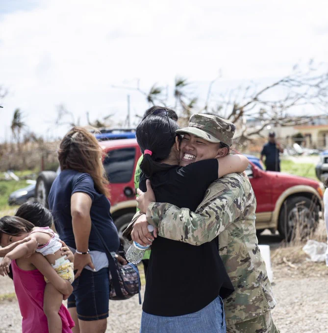 A member of the military hugs a disaster survivor in Saipan following Typhoon Yutu. In the background two children hug, and a woman surveys the damage.