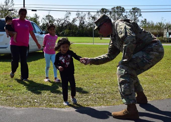 A member of the military smiles holds the hand of a skipping young girl as other members of her family walk behind her.