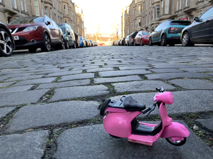 A little pink replica scooter in the middle of a sunny street paved with cobbles