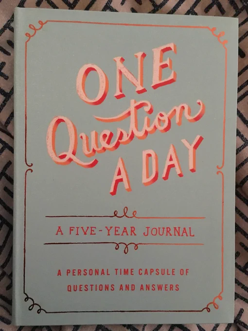 Image: book cover. It is duck egg blue, edged in gold, with curtly white and red writing. Text: One Question A Day: A Five Year Journal. A Personal Time Capsule Of Questions and Answers.