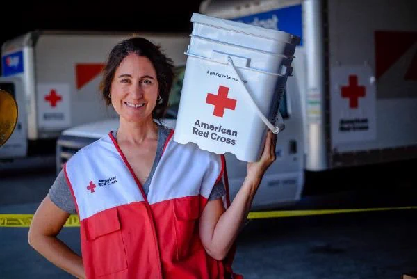 A volunteer for Red Cross wearing a uniform and posing with an American Red Cross bucket on her shoulder.