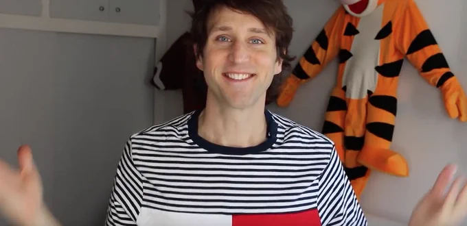 Josh Sundquist, comedian, and influencer, smiles at the camera wearing a Tommy Adaptive shirt as his iconic Tigger costume hangs in the background.
