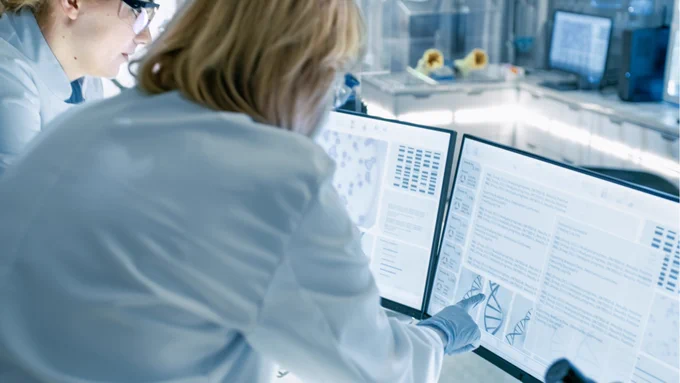 Two women in lab coats are examining images on a monitor. One is wearing gloves and points to a genome sequence on the screen.