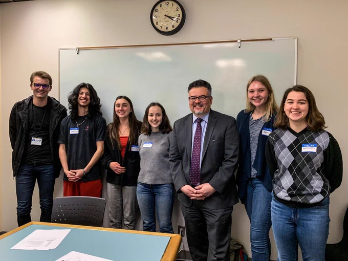 ‪6 students from Oregon Student Voice stand with Superintendent Guadalupe Guerrero in front of a white board with a clock above it.‬