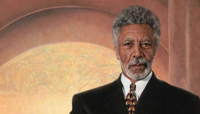 Ron Dellums, grey-haired, bearded, distinguished, standing against a frescoed wall