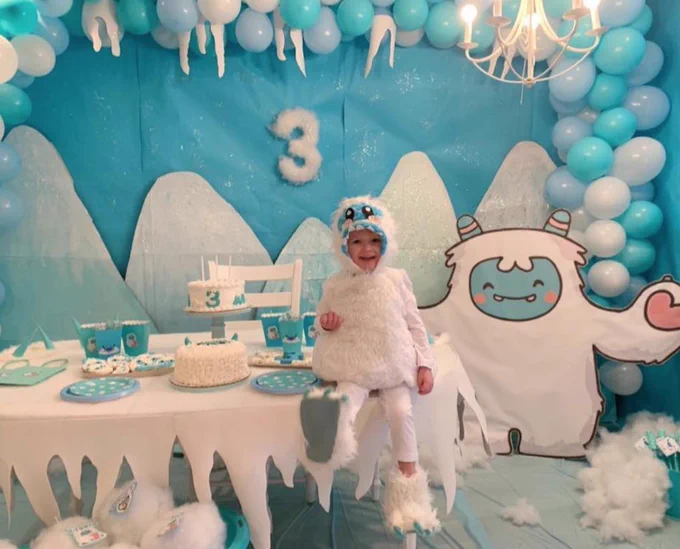 Holden, wearing a Yeti costume in front of a blue backdrop featuring mountains and a yeti cardboard cutout, smiling with a piece of cake