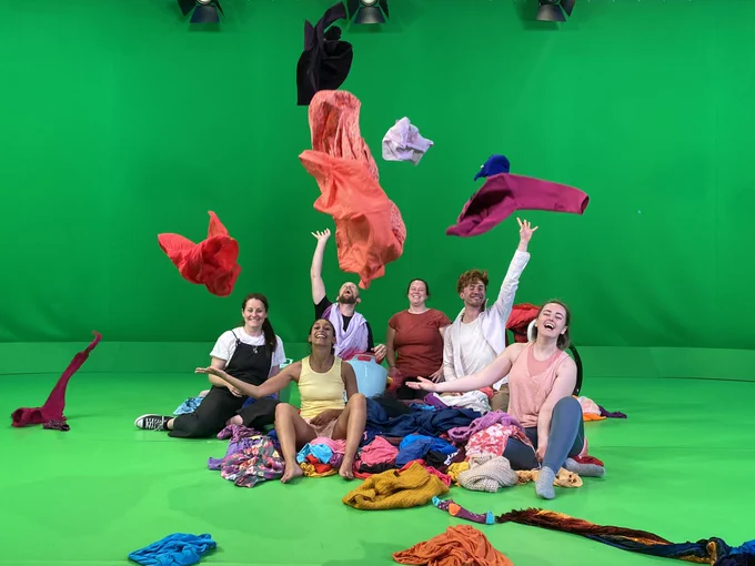 In a green room 6 adults sit ontop of a pile of clothes. Clothes hang in the air that they have just thrown above them.