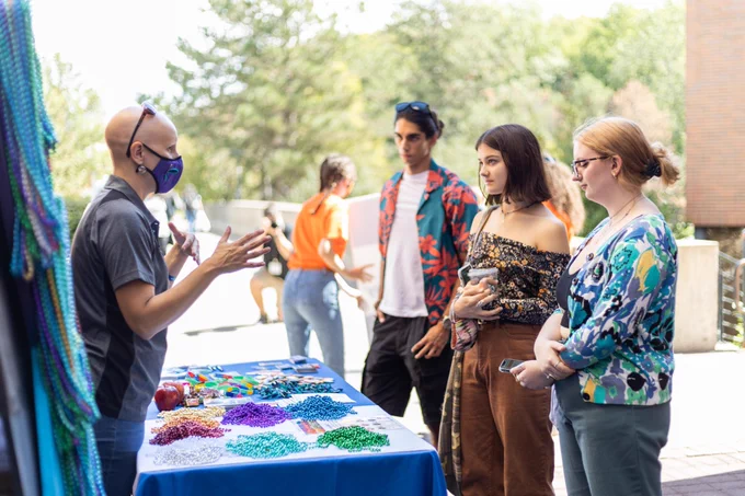 Students standing at table talking to staff member. Table full of colorful beaded necklaces.