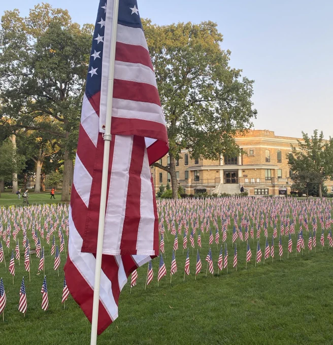 A large American flag with many small American flags in large numbers to honor those who died on September 11th. The flags are staked in green grass on the old portion of Bowling Green State University's campus, in front of a building of yellow brick.