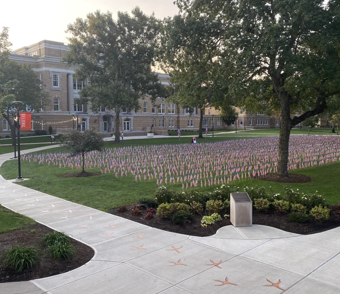 Many small American flags are staked in the ground to honor those who died on September 11th, 2001. The flags are in green grass on the old portion of Bowling Green State University's campus, in front of a building of yellow brick.