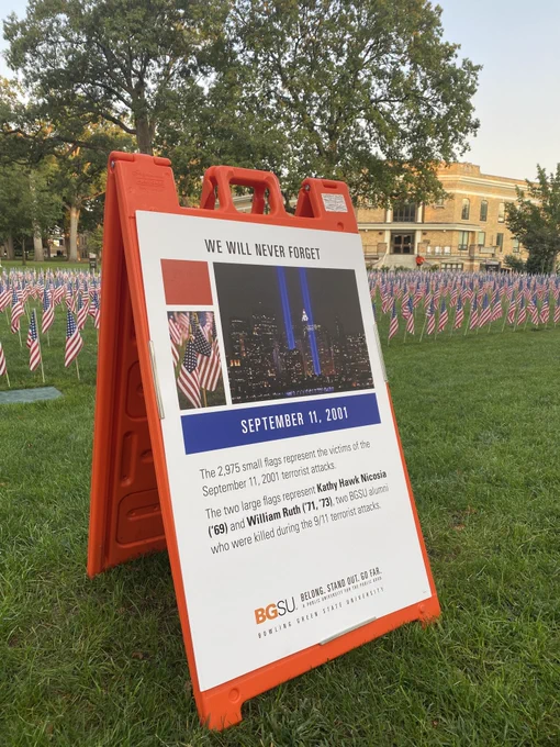 A board describing a field of American flags staked in the ground. The board pictures two blue lights rising out of a large scene of buildings that is downtown Manhattan. The rising blue lights signify where the twin towers---World Trade Center---were before they were destroyed on September 11th, 2001. The board with the picture reads: "The 2971 small flags represent the victims of the September 11, 2001 terrorist attacks. The two large flags [not pictured here] represent Kathy Hawk Nicosia ('69) and William Ruth ('71, '73), two BGSU alumni who were killed during the 9/11 terrorist attacks." The board and flags are staked in green grass on the old portion of Bowling Green State University's campus, in front of a building of yellow brick.