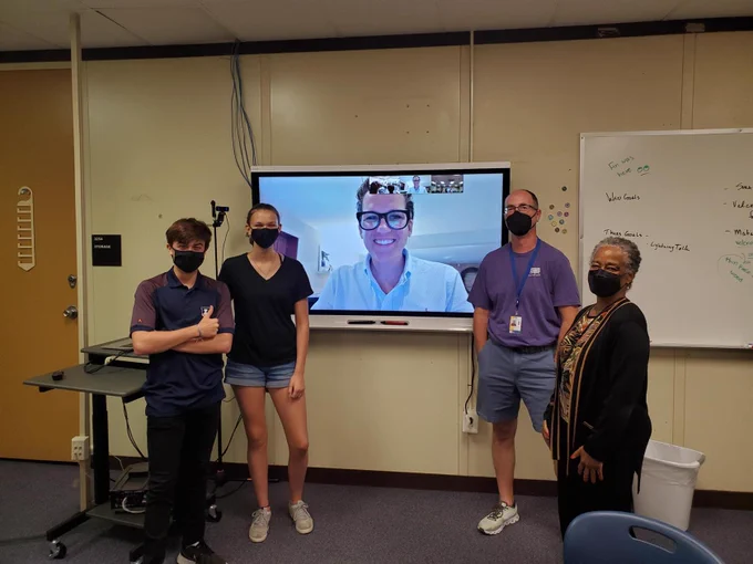 Four people, two high school students, two adults, stand in front of a monitor on which a fifth person is smiling. 