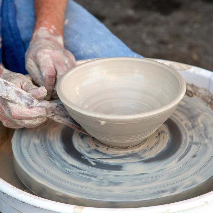 Photo of a pottery bowl being smoothed by a tool held by one hand, on a pottery wheel. The other hand rests beside it.