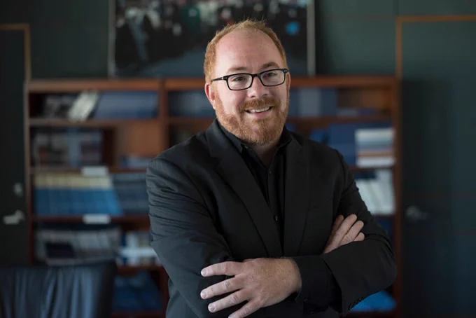 Dr. Matthew Kavanagh stands in the foreground with a bookshelf behind him.