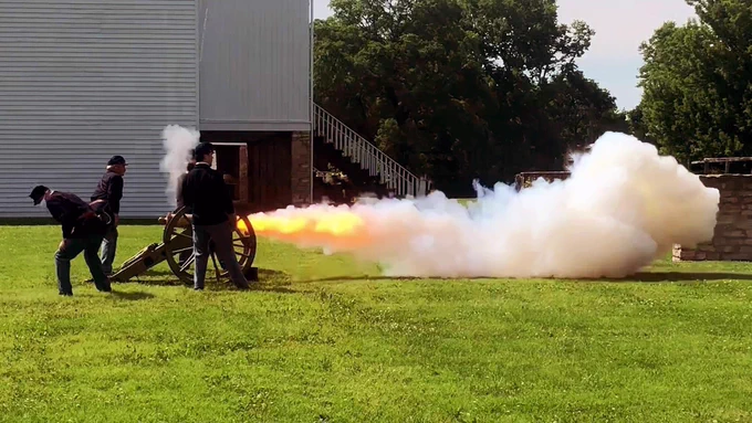 Four people dressed in dark blue civil war uniforms, firing a 12 pound mountain howitzer. Smoke and flames are coming from the barrel of the cannon.