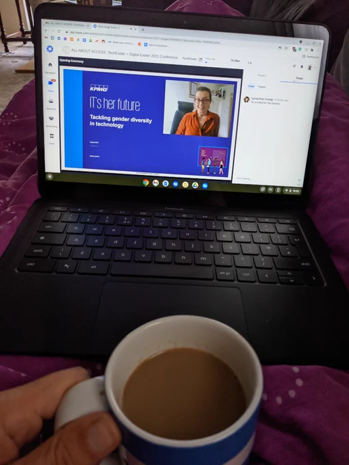 An open laptop with a conference talk showing sits on top of a quilted lap with a cup of coffee in front of it. The quilt it pink. The cup is blue and white.