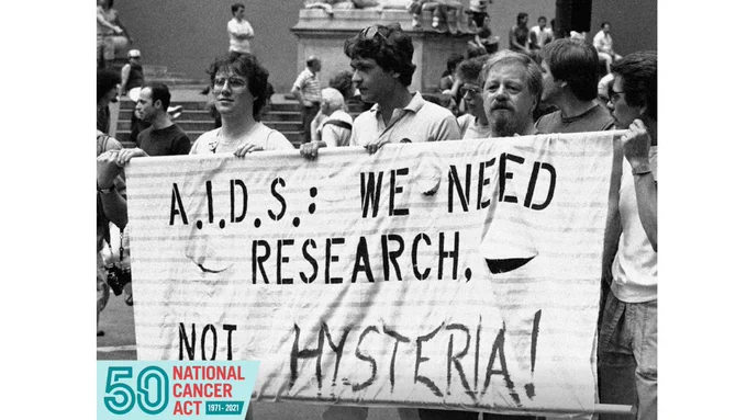 Three men hold a sign that reads, "A.I.D.S.: We Need Research. Not Hysteria!" In the background, there are various men milling about. There is a label added to the photo that says, "50 National Cancer Act 1971-2021." (National Cancer Institute).
